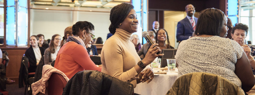 smiling woman holding a microphone and speaking from a table at a conference