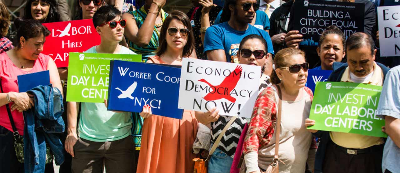 workers coop in nyc rally with signs that say "economic democracy now" and "invest in day laborer centers"